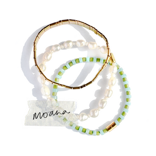 Three beaded stretch bracelets arranged on a white background: one gold-toned bracelet, one white pearl bracelet, and one mint green and gold-toned beaded bracelet. A small tag with the script brand name 'Mona' is placed near the bottom left.