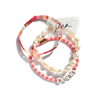 Three beaded bracelets with pink and coral beads, white baroque pearls, and a white letter bead spelling 'BUBS', arranged on a white background with a small paper tag reading 'pna'.