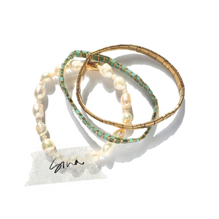 Three stacked bangles on a white background: two are gold-toned with turquoise and white pearl accents, and one is a gold-toned beaded bracelet. A small handwritten tag is attached.