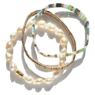 Three layered beaded bracelets on a white background. One bracelet features large cream-colored freshwater pearls, while the other two feature gold-plated tubes with white, light blue, and green beads.